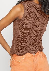 Brun ärmlös topp med en texturerad, rynkad design, som har en hög halsringning och slät tyg, tillsammans med orange byxor.