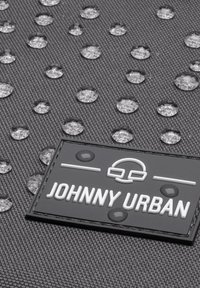 Material texturat negru cu picături de apă; o pată dreptunghiulară etichetată "JOHNNY URBAN" în alb și gri, cu accente circulare.