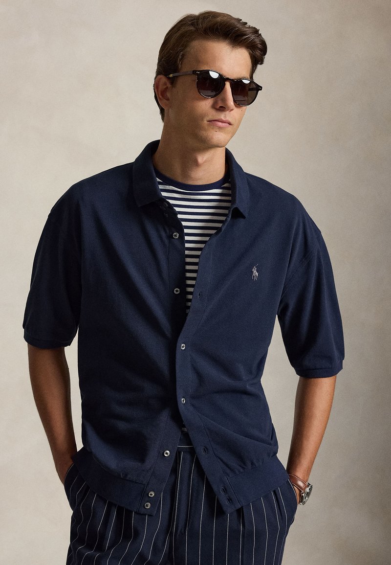 Άντρας που φοράει σκούρα γυαλιά ηλίου, μπλε ναυτικό πουκάμισο με κουμπιά πάνω από μια ριγέ μπλούζα και παντελόνι με λεπτές ρίγες, με τα χέρια στις τσέπες.