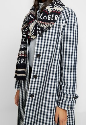 Personne portant un manteau à carreaux noir et blanc avec de gros boutons noirs et une écharpe à motifs noir, blanc et rouge enroulée autour du cou.