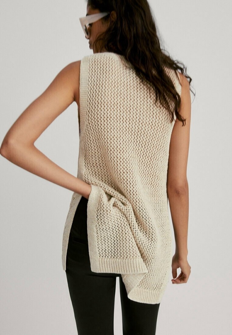 Femme aux longs cheveux portant un gilet en maille beige sans manches et un pantalon noir, ajustant le gilet par l'arrière avec la main.