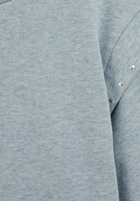 Svetlo modra majica s teksturirano tkanino. Ima kovinske žebljičke vzdolž ramenskega šiva. Zaobljen izrez in dolgi rokavi.