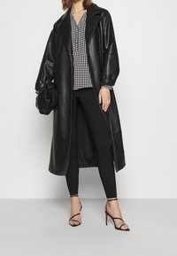 Sort læderlang jakke med drop-skuldre, kombineret med en bluse i houndstooth-mønster, sorte leggings og strap-heels i sort.