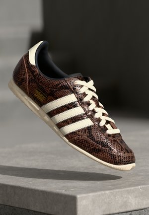 Brun struktureret Adidas sneaker med hvide striber og snørebånd ophængt over en betonkant i blødt lys.