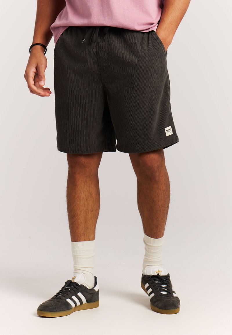 Uomo che indossa pantaloncini in velluto a coste neri, calzini bianchi, scarpe Adidas nere con strisce bianche e una maglietta rosa con le mani nelle tasche.