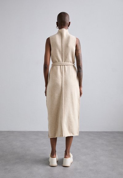 Abito midi in misto lana beige con design senza maniche, vita con cintura, fessure laterali e dettagli di cucitura sul retro. Indossato con sabot bianchi con tacco.