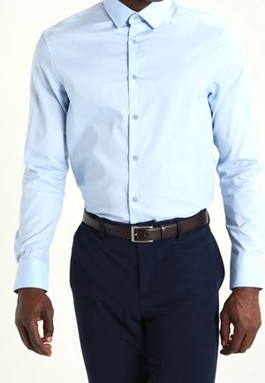 Camicia azzurro chiaro a maniche lunghe con colletto a punta, abbinata a pantaloni eleganti blu scuro e una cintura in pelle marrone.