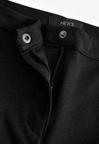 Pantalones de tela negra con cierre de cremallera y botón. Presentan una textura suave, una cintura plana y una etiqueta tejida que marca la marca "NEXT."