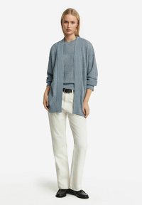 Ljusblå cardigan över en texturerad tröja, kombinerad med vita rakbeniga byxor och svarta loafers. Har ett öppet framstycke och uppvikta ärmar.