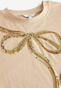 Beige T-shirt med en dekorativ design av paljetter i guld, grönt och koppar som är arrangerade i en rosettform med en slät tygtextur.