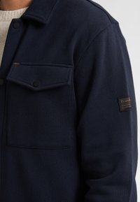 Marineblauwe fleecejack met twee borstzakken, zwarte drukknoopsluitingen en een gestructureerde stof. Voorzien van een merkpatch op de mouw.