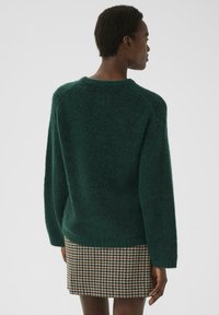 Vrouw in een donkergroene fuzzy trui en een bruin, zwart en crèmekleurig geruite rok, staande met de rug naar de camera tegen een effen achtergrond.