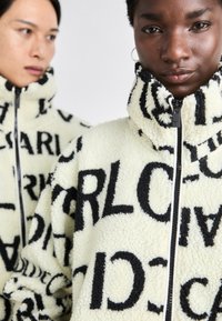 Veste en polaire crème avec de grands motifs textuels noirs. Elle présente un col haut et une fermeture éclair intégrale, mettant en valeur une surface douce et texturée.
