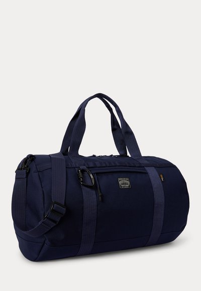 Ναυτικό μπλε σάκος duffel με διπλές λαβές μεταφοράς, ρυθμιζόμενο ιμάντα ώμου, μπροστινή τσέπη με φερμουάρ και μικρό επώνυμο ένθετο στο πλάι.