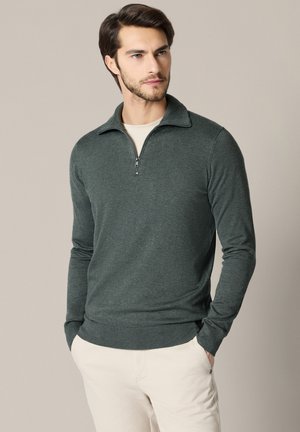 Mężczyzna w ciemnozielonym swetrze zapinanym do ćwiartki i jasnobeżowych spodniach, stojący z rękami w kieszeniach, lekko patrzący na bok.