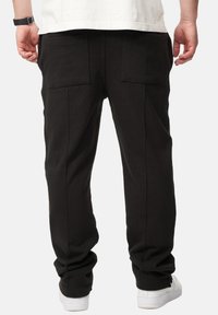 Schwarze Baumwollhosen mit lockerer Passform, ausgestattet mit zwei aufgesetzten Taschen auf der Rückseite und einem schmal zulaufenden Bein, kombiniert mit weißen Sneakern.