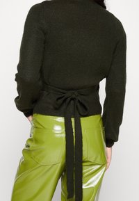 Dunkelgrüner gestrickter Pullover mit einer Detail-Schleife am Rücken, kombiniert mit glänzenden, hoch taillierten limettengrünen Kunstlederhosen mit Gesäßtaschen.