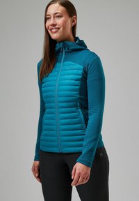 Teal hoodie met een gewatteerde voorkant, verticale geribbelde mouwen, volledige rits en een logo op de linkerborst. Gepaard met zwarte broek.