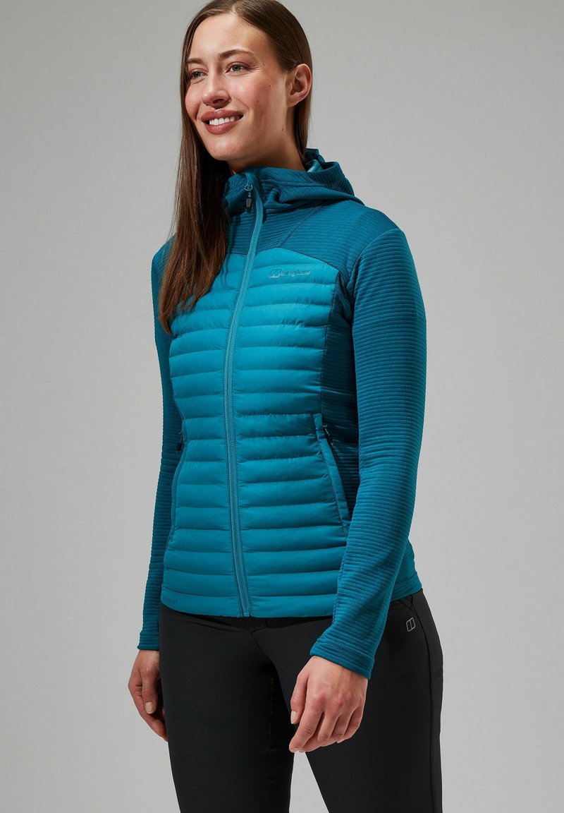 Teal hoodie met een gewatteerde voorkant, verticale geribbelde mouwen, volledige rits en een logo op de linkerborst. Gepaard met zwarte broek.