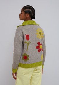 Ung kvinde med flettet hår iført en grå sweater med grøn kant og farverige blomsterprint, kombineret med lysegule bukser.