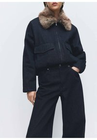 Tumma denimtakki tekoturkiskauluksella, jossa kaksi etutaskua ja vetoketjusuljenta, yhdistetty vastaaviin leveälahkeisiin farkkuihin.