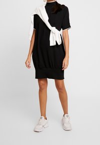 Černé krátké šaty s krátkými rukávy, volným střihem a nařaseným lemem. Přes ramena je přehozený bílý svetr. Kombinováno s bílými teniskami.