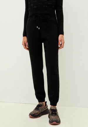 Personne portant un pantalon de jogging noir avec taille à cordon et des baskets noires avec des accents à imprimé léopard, debout devant un fond uni.