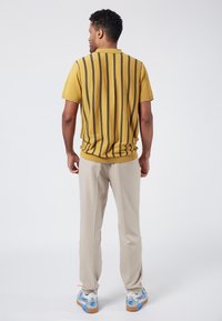 Gul stickad polo med vertikala svarta ränder, matchad med ljusbeige byxor. Modellen står med ryggen mot kameran, vilket visar detaljerna i outfiten.