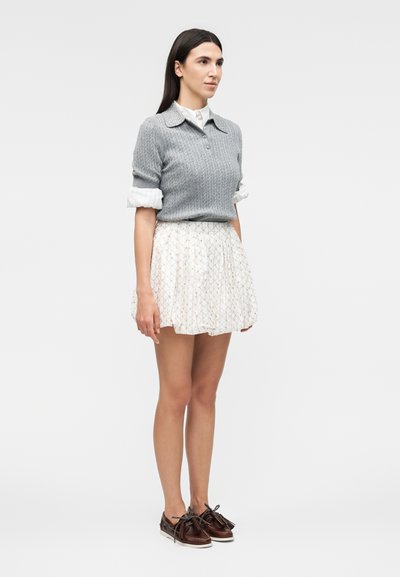 Γυναίκα που στέκεται, φορά γκρι πλεκτό πουλόβερ πάνω από λευκό πουκάμισο με γιακά, λευκή πλισέ φούστα και καφέ δερμάτινα μοκασίνια με φούντες.