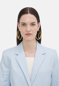 Cercei din aur în formă de cerc și o jachetă bleu deschis cu rever înclinat. Jacheta are un singur buzunar pe piept și un top de culoare crem dedesubt.