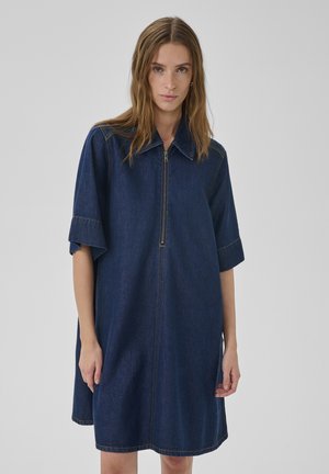 Femme portant une robe en jean bleu foncé à manches mi-longues et zipper frontal, debout devant un fond gris uni.