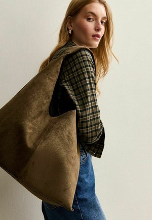 Blonde vrouw in een geruite shirt en blauwe jeans, met een grote, gestructureerde bruine tote bag over haar schouder, tegen een effen muur.