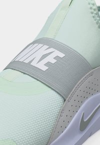 Κοντινή λήψη ενός ανοιχτό πράσινου αθλητικού παπουτσιού Nike με φαρδιά γκρι λουρακιό στο πάνω μέρος που φέρει το λευκό λογότυπο της Nike.