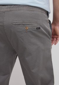 Pantaloni gri din bumbac cu detalii de buzunar cusute, accent din buton maro și un mic etichetă neagră pe spate. Țesătura are o textură netedă.