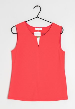 Růžový korálový top bez rukávů s texturou a mírným výstřihem do V, zavěšený na černém kovovém ramínku na jednobarevném pozadí.