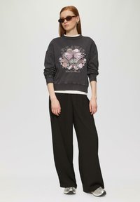 Szary sweter z graficznym motywem motyla i kwiatów, czarne spodnie o szerokich nogawkach oraz srebrne sneakersy. Model nosi nowoczesne okulary przeciwsłoneczne i warstwowy look.