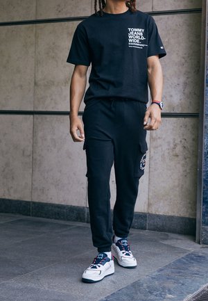 Black cotton t-shirt with white "Tommy Jeans" text. Black cargo pants with pockets. White sneakers featuring red and blue accents.