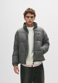 Grå pufferjakke med høj krave, lynlåslukning og quiltet mønster. Bæres over en hvid sweater og sorte jeans.