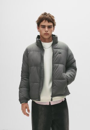 Grå pufferjakke med høj krave, lynlåslukning og quiltet mønster. Bæres over en hvid sweater og sorte jeans.