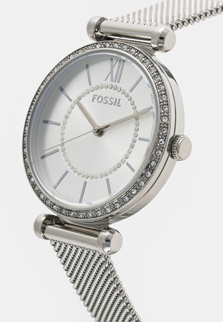 Reloj con correa de malla plateada, caja redonda, esfera blanca, números romanos y adornos de cristal alrededor del bisel. Marca: Fossil.