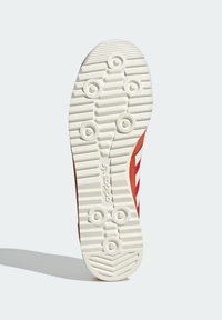 Röd sneaker sula med en texturerad yta, som har cirkulära och linjära mönster för grepp, samt en vit mellansula med en varumärkeslogotyp.