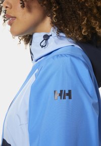 Blauwe waterdichte jas met een hoge kraag, verstelbare trekkoord en een zwart logo-detail op de mouw. Zachte textuur.