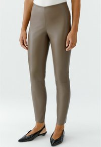Personne portant des leggings en simili cuir taupe et des escarpins noirs à bout pointu, se tenant contre un fond clair et uni.