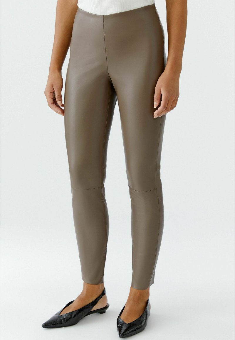 Person, die taupefarbene Kunstlederleggings und schwarze spitze Slingback-Flats trägt, steht vor einem schlichten, hellen Hintergrund.