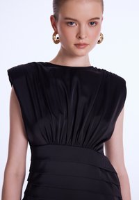 Robe noire avec un corsage structuré et plissé, un col large et une jupe à plusieurs couches. Texture en satin avec un subtil éclat. Boucles d'oreilles en or en forme de cercle.