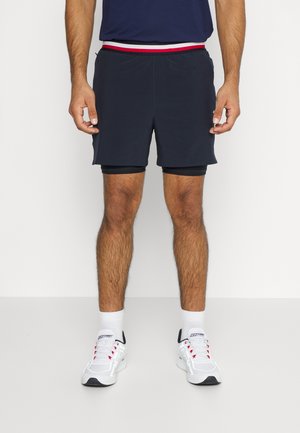 Mężczyzna w granatowych krótkich spodenkach sportowych z wbudowanymi czarnymi spodniami kompresyjnymi, białych skarpetkach i białych tenisówkach z czerwonymi sznurówkami na jednolitym tle.