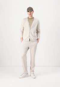 Cremefarbener Cardigan mit Knöpfen, olivfarbenes T-Shirt, hellbeige Hose und weiße Sneakers. Der Hut passt zur Farbe des Shirts.