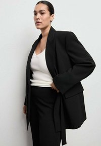 Blazer negro entallado con mangas anchas, detalle de bolsillo y cintura con cinturón, llevado sobre una camiseta blanca y pantalones negros. Textura suave.