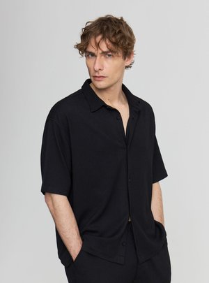 Giovane uomo dai capelli ricci che indossa una camicia nera a maniche corte larga e pantaloni coordinati, in piedi con le mani nelle tasche, guarda davanti a sé.