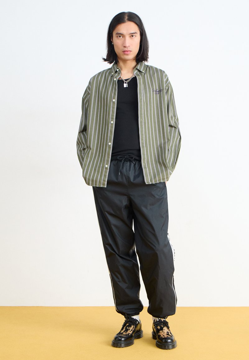 Chemise à boutons rayée verte et blanche, débardeur noir, pantalon noir à cordon de serrage et chaussures à motif. Design simple, coupe décontractée.